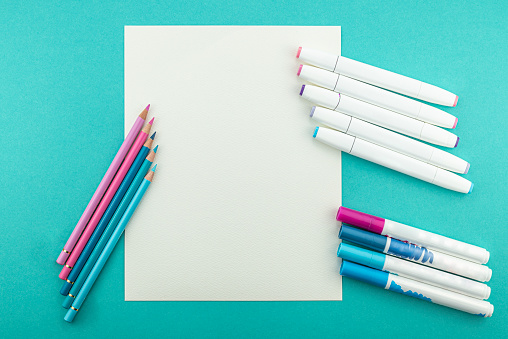 Overhead studio image of a blank paper on turquoise blue background. Around the paper there are color pencils and different kinds of pens, all in blue and pink tones.