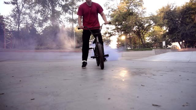 Bmx rider practicing in the empty skate park, riding on the bike while blue smoke bomb is burning on bike's back wheel. Man stops strickly before the camera, close up of rhe wheel