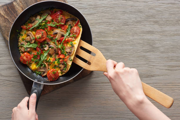 cropped view of woman cooking omelet with mushrooms, tomatoes and greens on frying pan with wooden shovel cropped view of woman cooking omelet with mushrooms, tomatoes and greens on frying pan with wooden shovel cooking pan overhead stock pictures, royalty-free photos & images