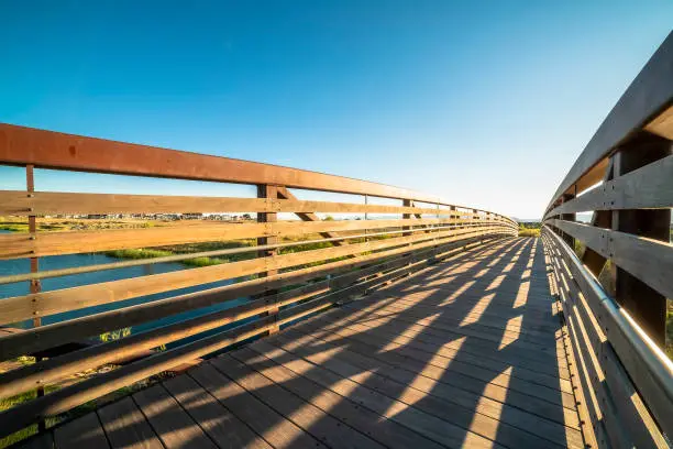 View from of the wooden deck and metal guardrails of a bridge on a sunny day. Scenic landscape with lake and homes can also be seen under the blue sky.