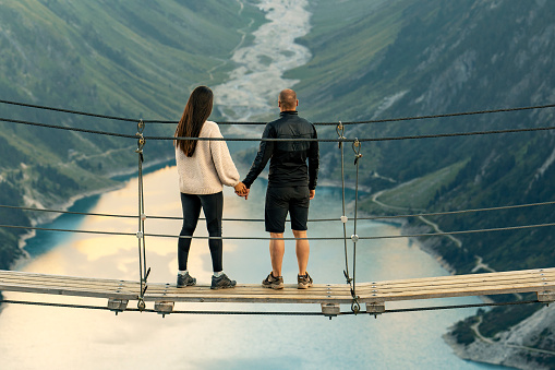 Happy couple on holiday. Mountain landscape. Boyfriend and girlfriend. Romantic moment after proposal or engagement. Passionate lovers on summer vacation. Couple in love enjoying the view from Schlegeis Stausee (Schlegeis Lake) in Tyrol, Austria. Man standing with his girlfriend at rope bridge, holding hands at Schlegeis glacier and beautiful blue lake in the mountains of Tirol, Austria.