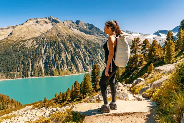 Beautiful alpine landscape with young woman standing with backpack looking at azure mountain lake in the background, Zillertal Alps, Austria