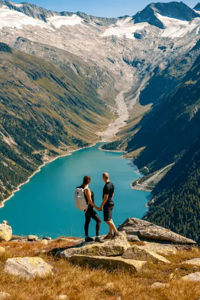 Beautiful alpine landscape with young couple holding hands, man and woman in backpack standing, looking at azure mountain lake in the background, Zillertal Alps, Austria