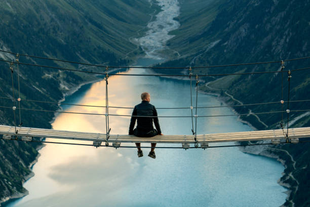 This is a stunning location! Rear view of Man sitting on rope bridge looking at Mountain landscape. Lonely man enjoying the view from Schlegeis Stausee (Schlegeis Lake) in Tyrol, Austria. Man looking at Schlegeis glacier and beautiful blue lake in the mountains of Tirol, Austria. wilderness photos stock pictures, royalty-free photos & images