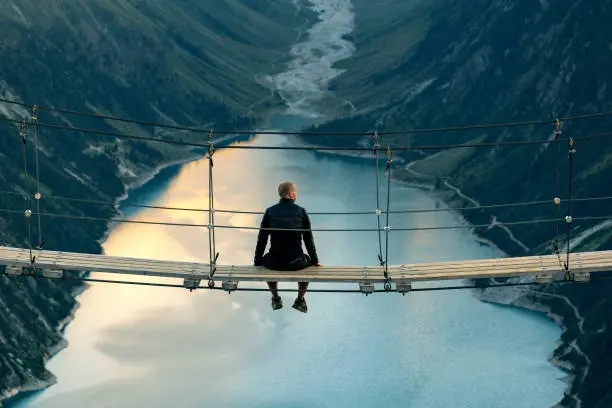 Rear view of Man sitting on rope bridge looking at Mountain landscape. Lonely man enjoying the view from Schlegeis Stausee (Schlegeis Lake) in Tyrol, Austria. Man looking at Schlegeis glacier and beautiful blue lake in the mountains of Tirol, Austria.