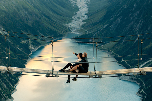 Happy couple on holiday. Mountain landscape. Boyfriend sitting with his girlfriend on rope brige. Romantic moment after proposal or engagement. Passionate lovers on summer vacation. Couple in love enjoying the view from Schlegeis Stausee (Schlegeis Lake) in Tyrol, Austria. Man and his girlfriend at Schlegeis glacier and beautiful blue lake in the mountains of Tirol, Austria.