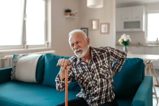 Elderly Man suffers from Back Pain Senior man suffers from back pain. He sits on the sofa in the living room in one hand holding a walking stick while his other hand is on his painful back with a painful grimace on his face. tetanospasmin stock pictures, royalty-free photos & images