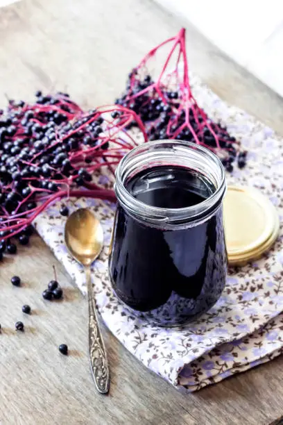 Homemade black elderberry syrup in glass jar and bunches of black elderberry in background