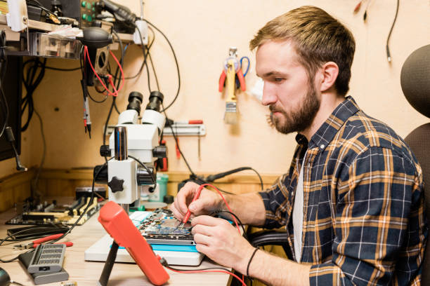 istock Young bearded mechanic with two soldering-irons sitting by workplace in workshop 1184924445