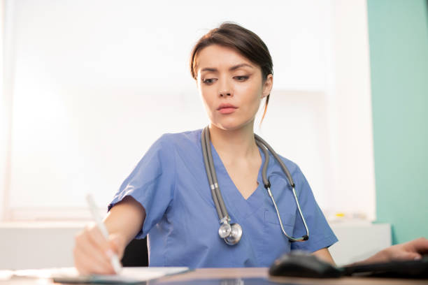 istock Young serious doctor or intern in blue uniform making working notes by workplace 1184925857