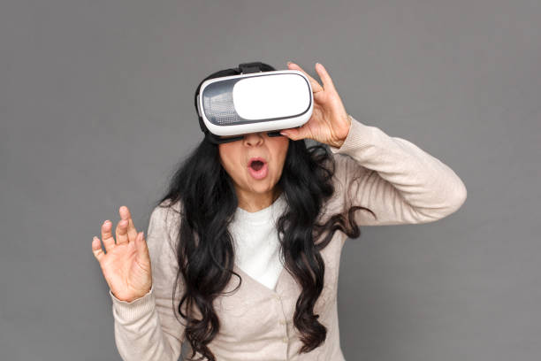 istock Freestyle. Mature lady in virtual reality headset standing isolated on grey playing surprised 1184935363