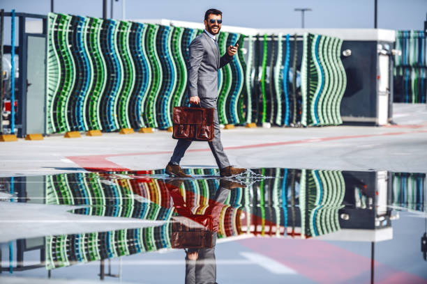 full length of handsome caucasian bearded businessman in suit and with sunglasses holding briefcase and smart phone while crossing over the puddle. - wet spy men suit imagens e fotografias de stock