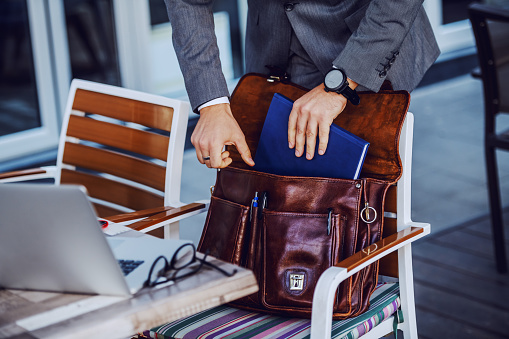 Cropped picture of elegant businessman in suit taking out agenda from his leather bag. Cafe exterior.