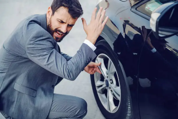 Photo of Angry handsome Caucasian businessman crouching next to his car, yelling and swearing. Flat tire.