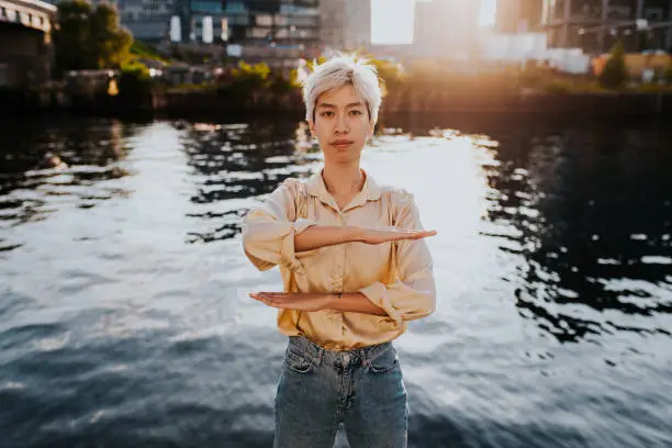 Photo of Portrait Of Young Woman Gesturing Equal Sign While Standing Against Lake In City