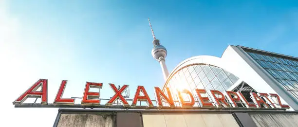 Wide angle panorama of Alexanderplatz sign at railway and subway station with tv tower behind - Side view of world famous landmark in Berlin capital of Germany - Warm vivid sunshine filter