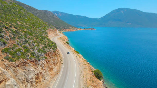 DRONE: Flying behind a tourist car exploring the remote island in the Mediterranean by driving down a scenic coastal road. Tourists cruising along the breathtaking shoreline of Lefkada in a grey car.