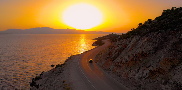 AERIAL: Black SUV drives up a winding asphalt road with a perfect view of the calm ocean at sunrise. Tourists on relaxing summer vacation in Lefkada exploring the famous island at colorful sunset.