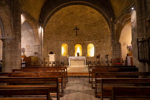Bright yellow lights illuminate the inside of an old Catholic church in France. Empty aisle leads past wooden benches and towards the medieval stone altar covered by a white tablecloth with ornaments.