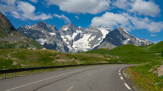 Spectacular snowy Alps tower high above the idyllic countryside in France. Empty asphalt road runs past the vibrant meadows and breathtaking mountain range. Stunning views from the grand alpine route