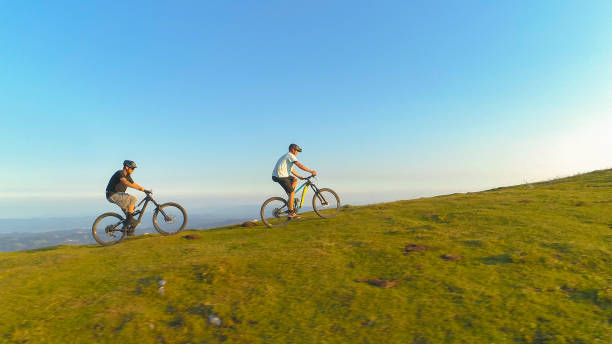 istock DRONE: Two mountain bikers ride their electric bicycles up a steep grassy hill. 1185005721
