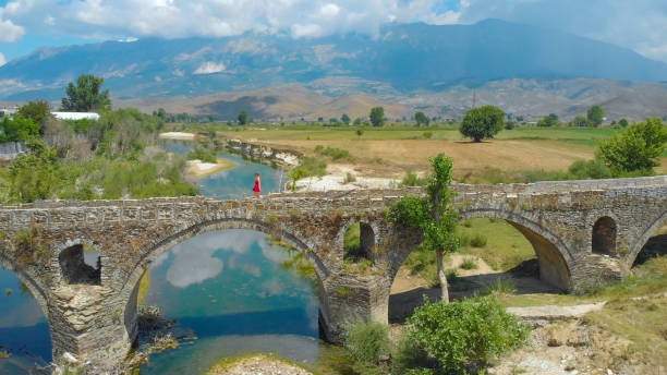 AERIAL: Woman in bright red dress strolling along the decaying arched bridge crossing the stream drying up in the hot countryside in the Balkan. Tourist girl enjoying a stroll down the ottoman bridge.