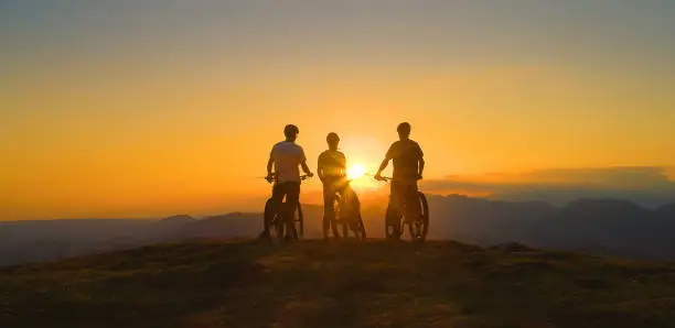 LOW ANGLE, SILHOUETTE, SUN FLARE: Golden evening sun rays illuminate cross country bikers on the mountaintop observing the scenic landscape. Three friends watch sunset after bicycle ride in mountains