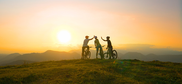 SUN FLARE: Mountain biking friends high five after reaching the scenic summit at breathtaking golden sunrise. Cross country cyclists celebrate a successful mountain biking adventure on a sunny evening