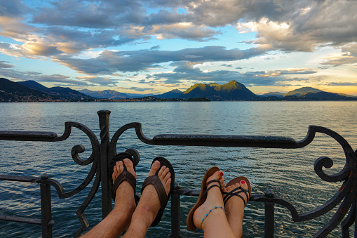 CLOSE UP: Tourist couple rests their feet on the metal railing while observing the breathtaking lake on a picturesque summer evening. Young man and woman wearing flip flops relax by Lago di Maggiore.