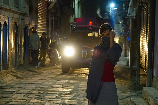 PORTRAIT: Young woman covers her face with a gray scarf while walking down the dark street in Kathmandu. Large car illuminates female tourist exploring the streets of Kathmandu in middle of the night.