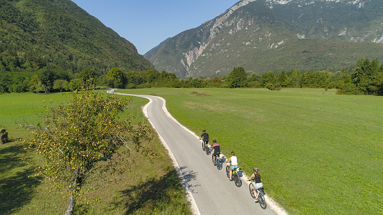 AERIAL: A group of tourists ride electric bicycles along the empty asphalt road in the scenic green valley in Slovenian countryside. Mountain bikers cruise through the picturesque nature on sunny day.