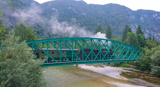 Retired locomotive chugs across the river running through the green mountains in Bohinj. Black vintage steam engine takes a group of tourists on a sightseeing trip of beautiful mountains in Slovenia