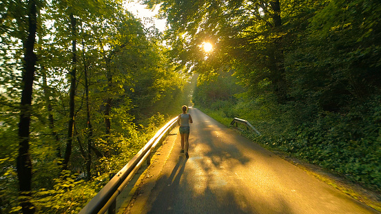 AERIAL, LENS FLARE: Unrecognizable young woman runs down an empty asphalt road leading through the calm woods. Flying behind unrecognizable fit girl exercising in nature on a beautiful spring morning.