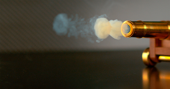 MACRO, DOF: A small cloud of smoke rolls out of the barrel of a miniature cannon after firing a round shot across the room. Vintage brass cannon sitting on shiny surface about to shoot a cannonball.