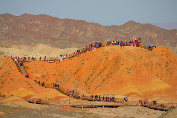 DRONE: Tourists move along the walkways leading up a hill in Danxia landform. DRONE: Masses of Asian tourists move along the walkways leading up a rocky hill in the Zhangye Danxia landform. Travelers walk along path leading up to hilltop offering a view of Rainbow mountains. danxia landform stock pictures, royalty-free photos & images