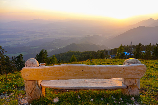 CLOSE UP: Golden evening sunbeams shine on empty wooden bench in the mountains overlooking the breathtaking green valley. Panoramic view of the endless evening lit countryside spans below the bench.