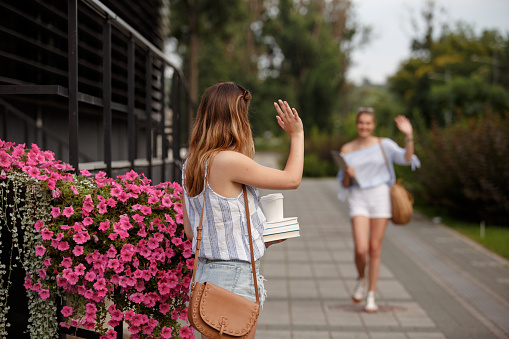 Shot of young woman standing on the street and waving to a friend that is approaching her to meet for a coffee and a relaxing walk together.
