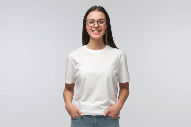 주머니에 손을 들고 서있는 젊은 웃는 여자는 회색 배경에 고립 된 복사 공간이있는 빈 흰색 티셔츠를 입고 있습니다. - 여자 이미지 뉴스 사진 이미지