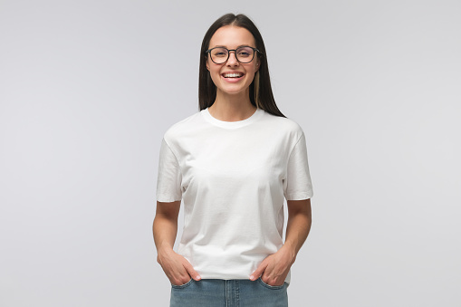 Young laughing woman standing with hands in pockets, wearing blank white t-shirt with copy space, isolated on gray background