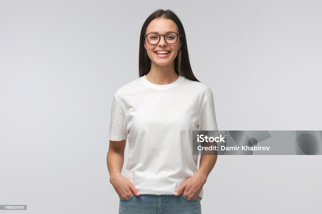 Young laughing woman standing with hands in pockets, wearing blank white t-shirt with copy space, isolated on gray background Women Stock Photo