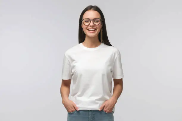 Photo of Young laughing woman standing with hands in pockets, wearing blank white t-shirt with copy space, isolated on gray background