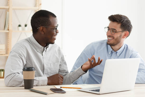 istock Teamwork concept. Diverse smiling modern businessmen using laptop together, african coworker showing something with hand to his caucasian colleague at white office 1185028197