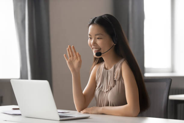 Smiling asian businesswoman wearing wireless headset waving doing video chat Smiling asian businesswoman wearing wireless headset waving hand doing video chat looking at laptop consulting customer sitting at workplace, female call center service support agent talking on skype school receptionist stock pictures, royalty-free photos & images