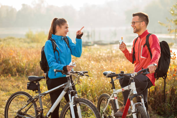 Playful happy young couple eating energy bars while taking a break from an early morning bicycle ride by the lake or river. Playful happy young couple eating energy bars while taking a break from an early morning bicycle ride by the lake or river. Active and healthy lifestyle red white blue bike stock pictures, royalty-free photos & images
