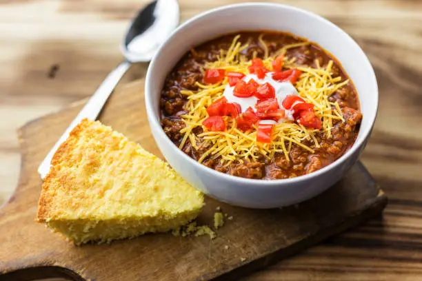Cornbread and chili, on a wood background with room for copy. Tomatoes, cheese and sour cream toppings are on the chili.