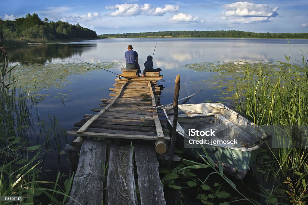 Fisherman on the lake A fisherman sits at the end of a dock with his fishing pole in hand.  A small boat is next to the dock, and water plants grow around it. Fishing Stock Photo