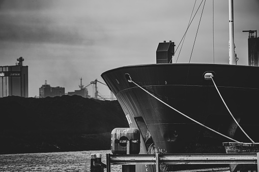The bow of a cargo ship docked at port, near by a mound of industrial waste and silos.  Belfast, Northern Ireland.  Monochrome.