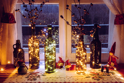 Cute and warm Christmas decoration set with vintage beer bottles and wine bottles filled with micro led party lights, behind the window is countryside forest. Bottles vases for red artificial berry.