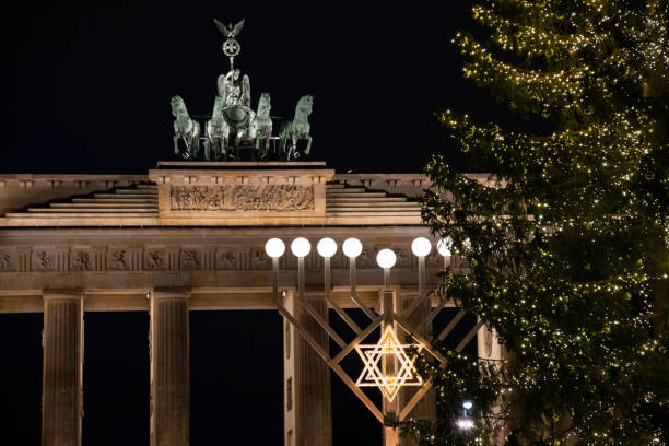 Religious symbol and shiny Christmas tree in front of Brandenburg Gate neoclassical monument in Berlin Germany. Night cityscape with Jewish Hanukah decorations and traditional Christmas decorations stock photo