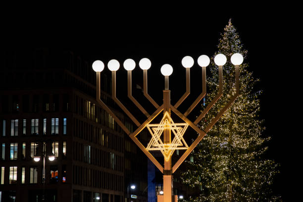 Huge illuminated menorah with round lamps and lighted Jewish symbol David's star on blurry background of Christmas tree in Berlin Germany. Night cityscape with Hanukah decorations stock photo